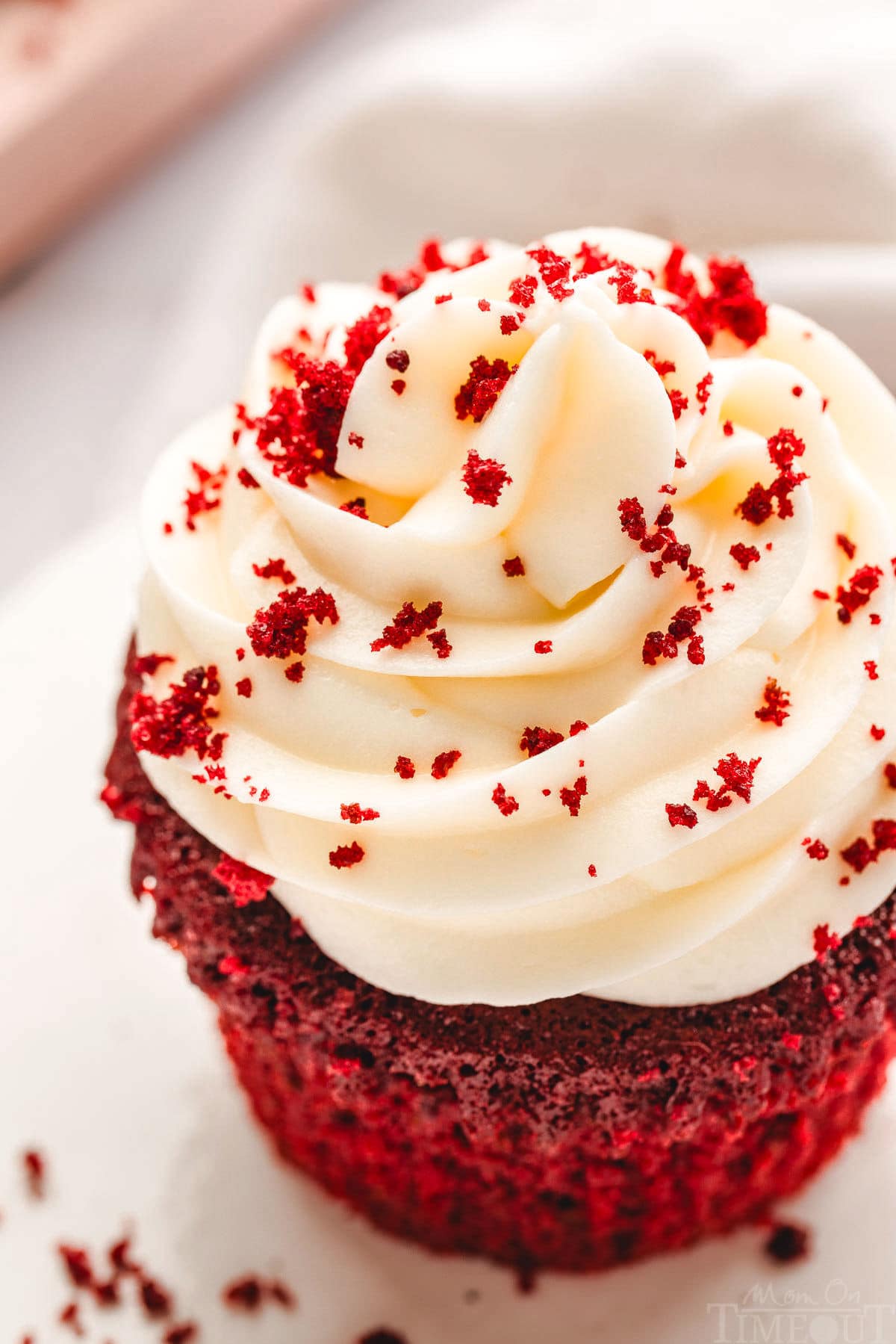 top down close up look at creamy creamy cheese frosting on a red velvet cupcake on white plate. red velvet crumbs have been sprinkled on top of the frosting.