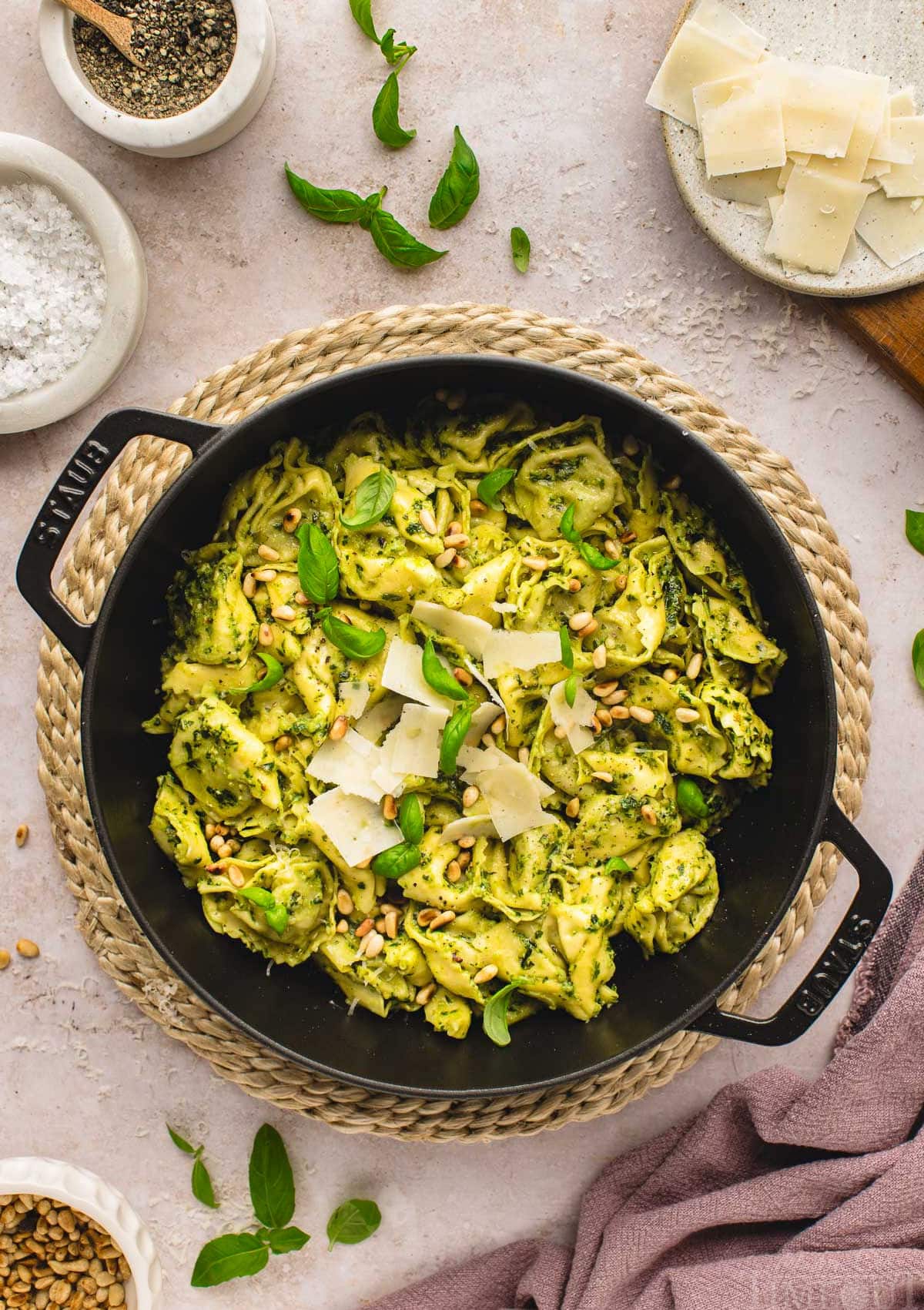 top down shot of pesto tortellini recipe in large black cast iron skillet topped with shaved parmesan, toasted pine nuts and fresh basil leaves. small bowls of parmesan, salt and pepper can be seen around the skillet.