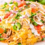 round white plate loaded high with pulled pork nachos topped with sour cream, jalapenos, tomatoes and red onions. a whole tomato and cilantro can be seen in the background.