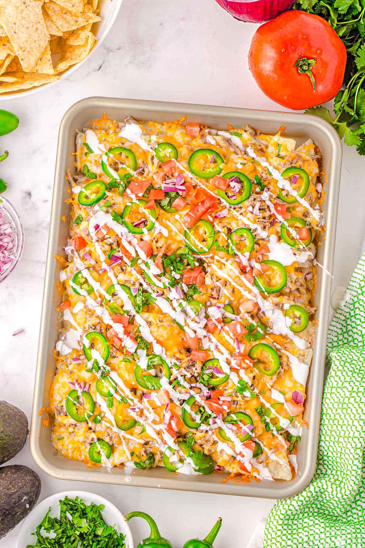 top down view of large baking sheet filled with loaded pulled pork nachos with a sour cream drizzle, sliced jalapenos, tomatoes, cilantro, and red onions. Ready to be enjoyed.