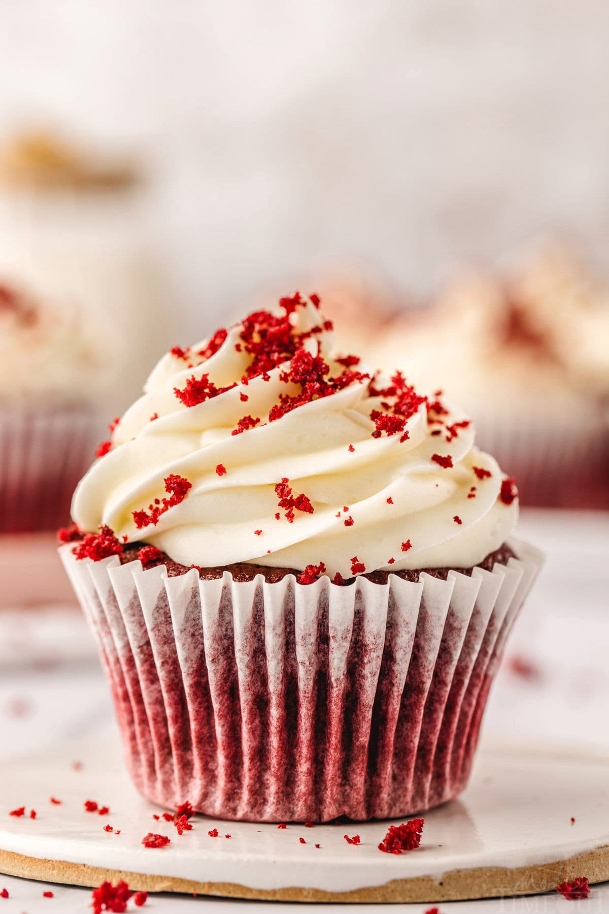 red velvet cupcake in a white liner sitting on white pottery plate topped with velvety cream cheese frosting and sprinkle with red velvet crumbs. more cupcakes can be seen in the background.