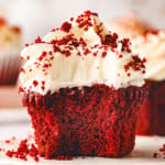 red velvet cupcake sitting on white pottery plate topped with velvety cream cheese frosting and sprinkle with red velvet crumbs.