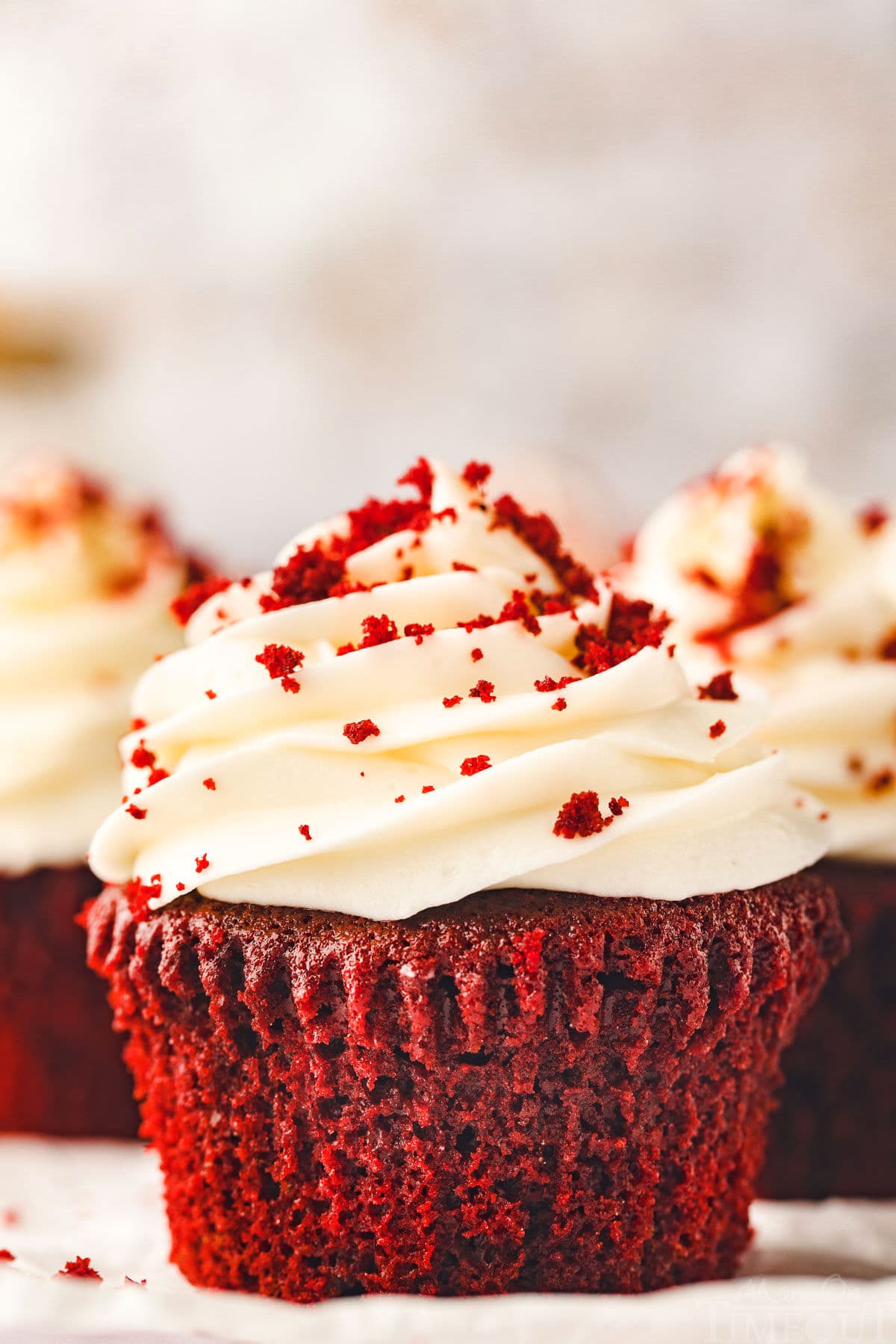red velvet cupcake sitting on white pottery plate topped with velvety cream cheese frosting and sprinkle with red velvet crumbs. more cupcakes can be seen in the background.