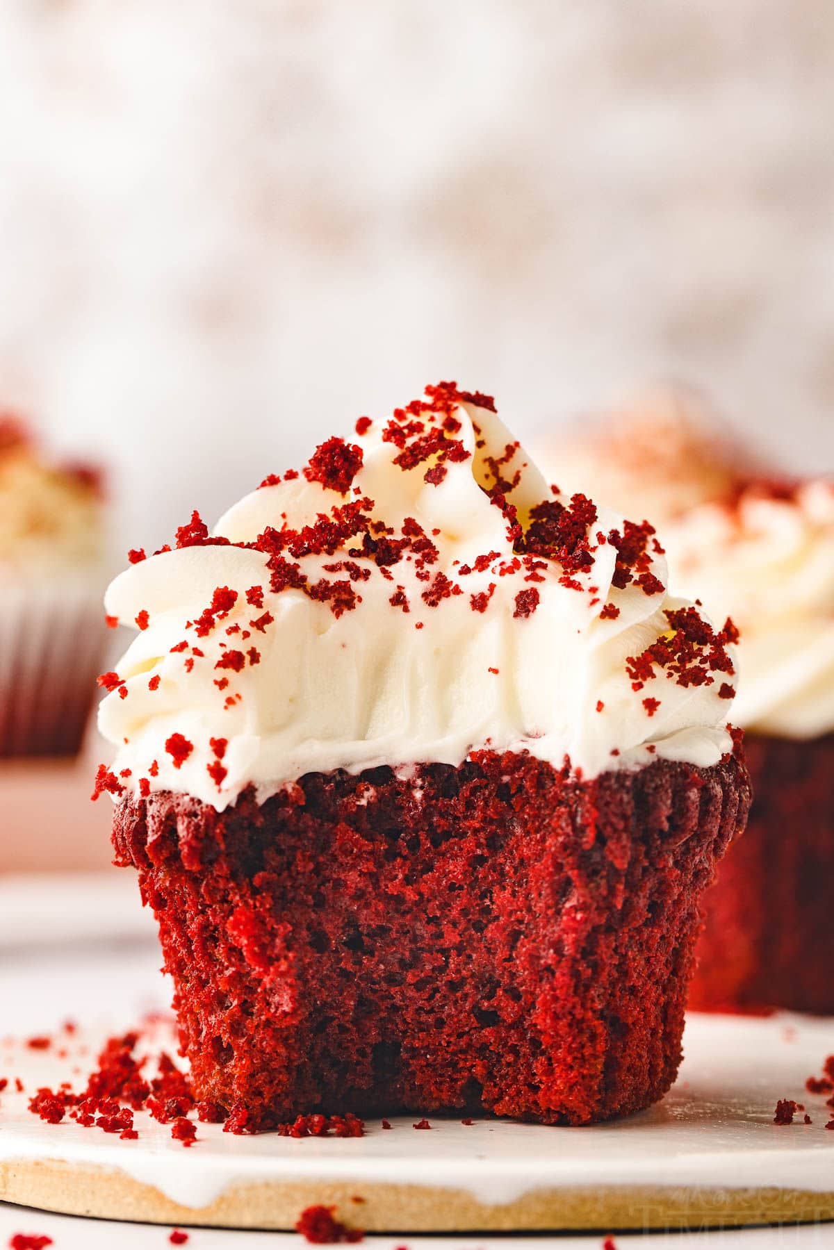 red velvet cupcake sitting on white pottery plate topped with velvety cream cheese frosting and sprinkle with red velvet crumbs. 