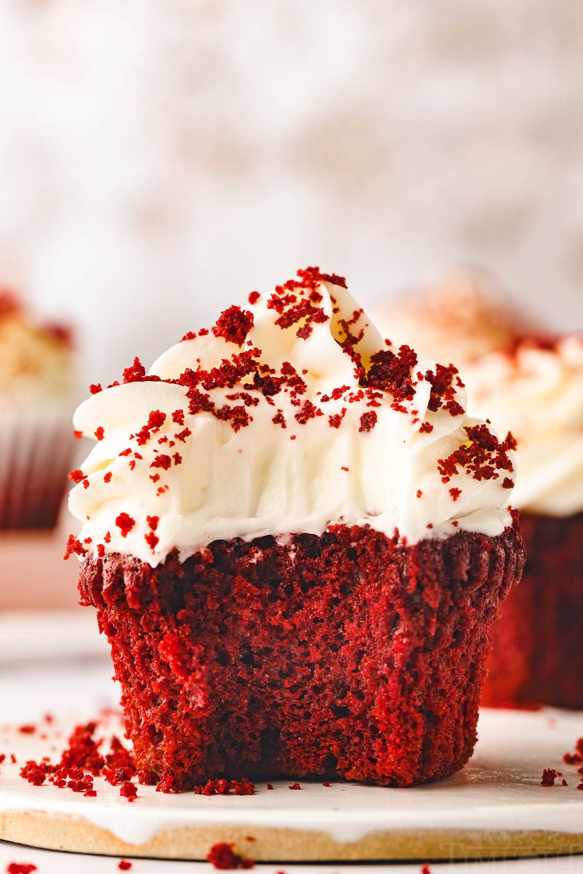 red velvet cupcake sitting on white pottery plate topped with velvety cream cheese frosting and sprinkle with red velvet crumbs.