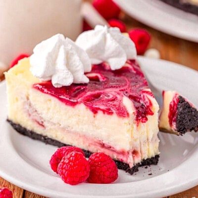 slice of white chocolate raspberry cheesecake on a round white plate topped with whipped cream. three raspberries are sitting next to the cheesecake on the plate.