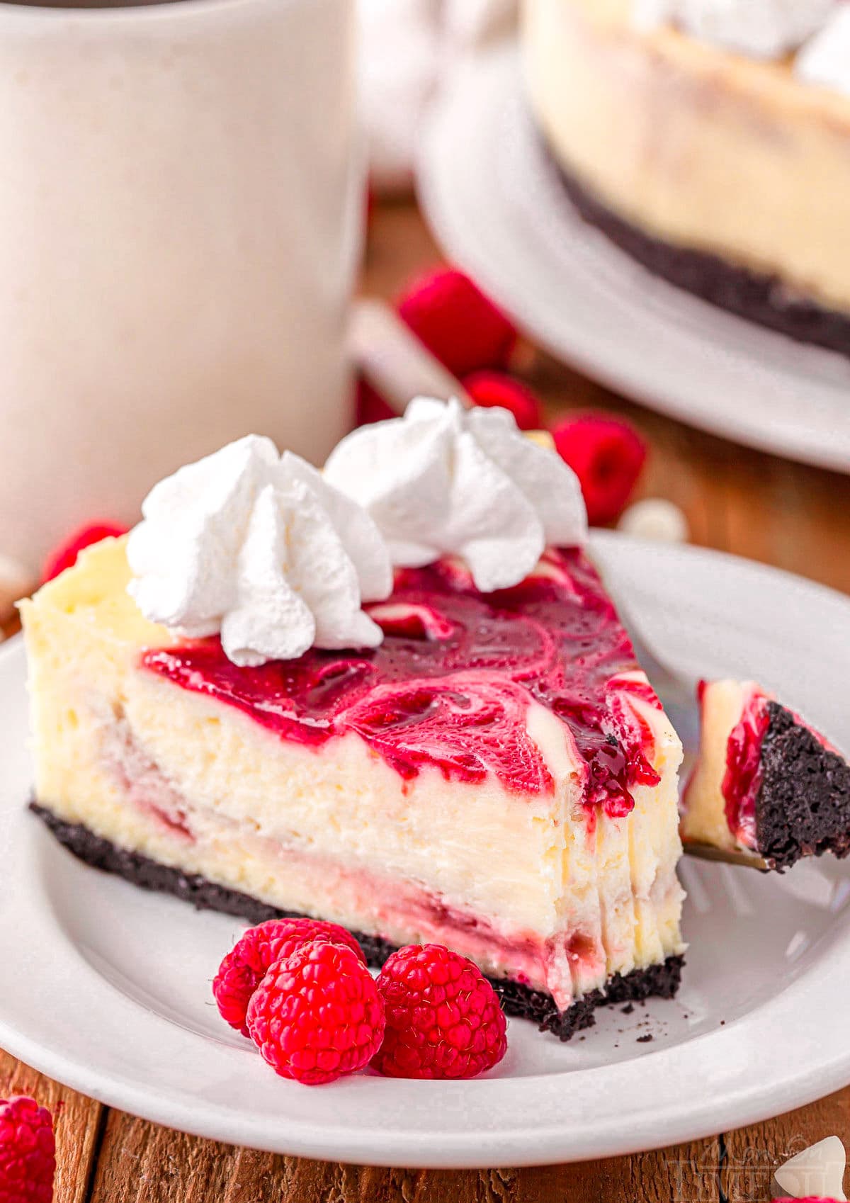 slice of white chocolate raspberry cheesecake on a round white plate topped with whipped cream. three raspberries are sitting next to the cheesecake on the plate.