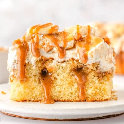 piece of banoffee poke cake on a white plate. the cake has been topped with whipped cream, bananas and dulce de leche sauce.