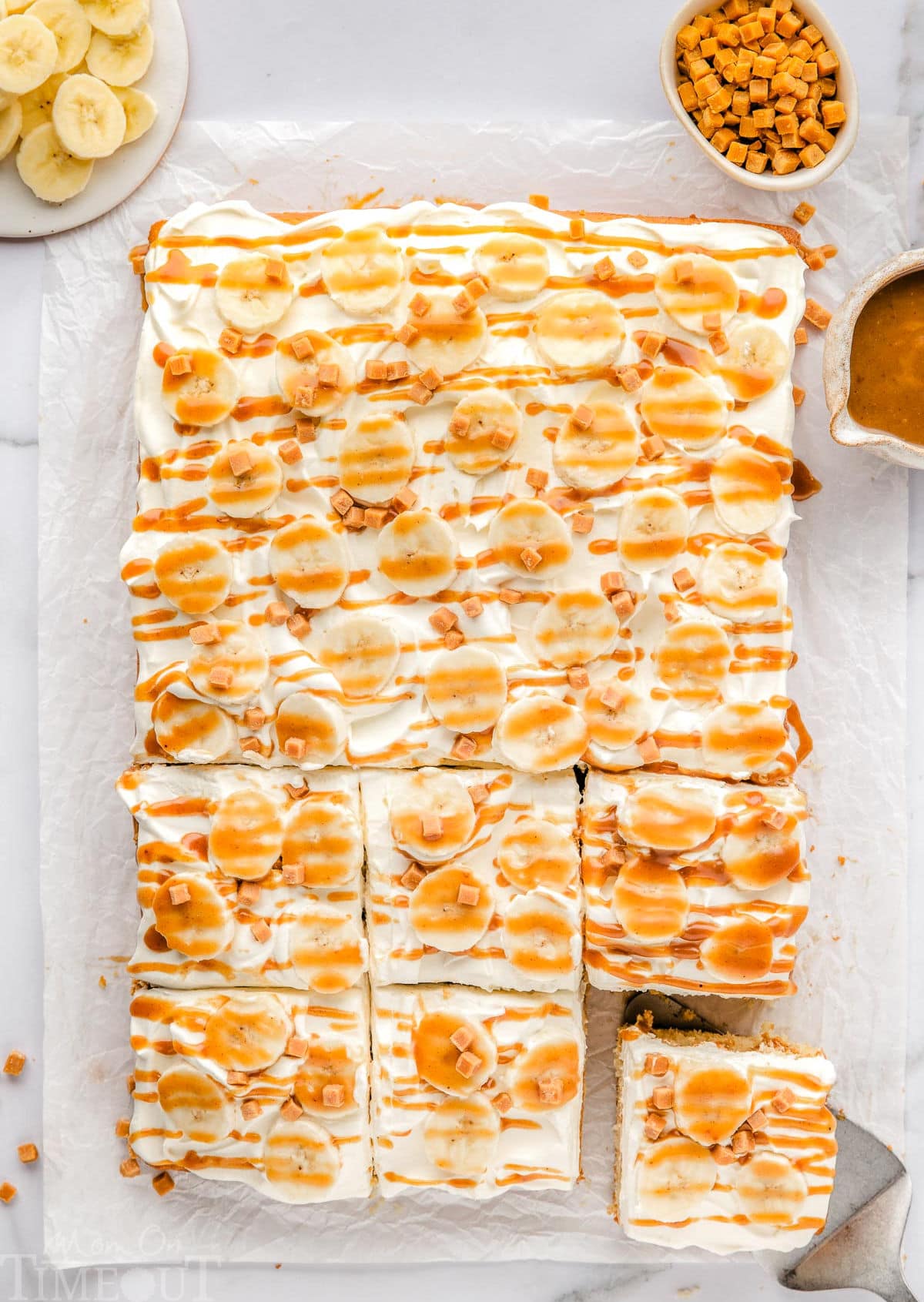 top down view of banoffee poke cake with six pieces cut. cake is sitting on white parchment. bowl of slices bananas and toffee bits are seen next to the cake.