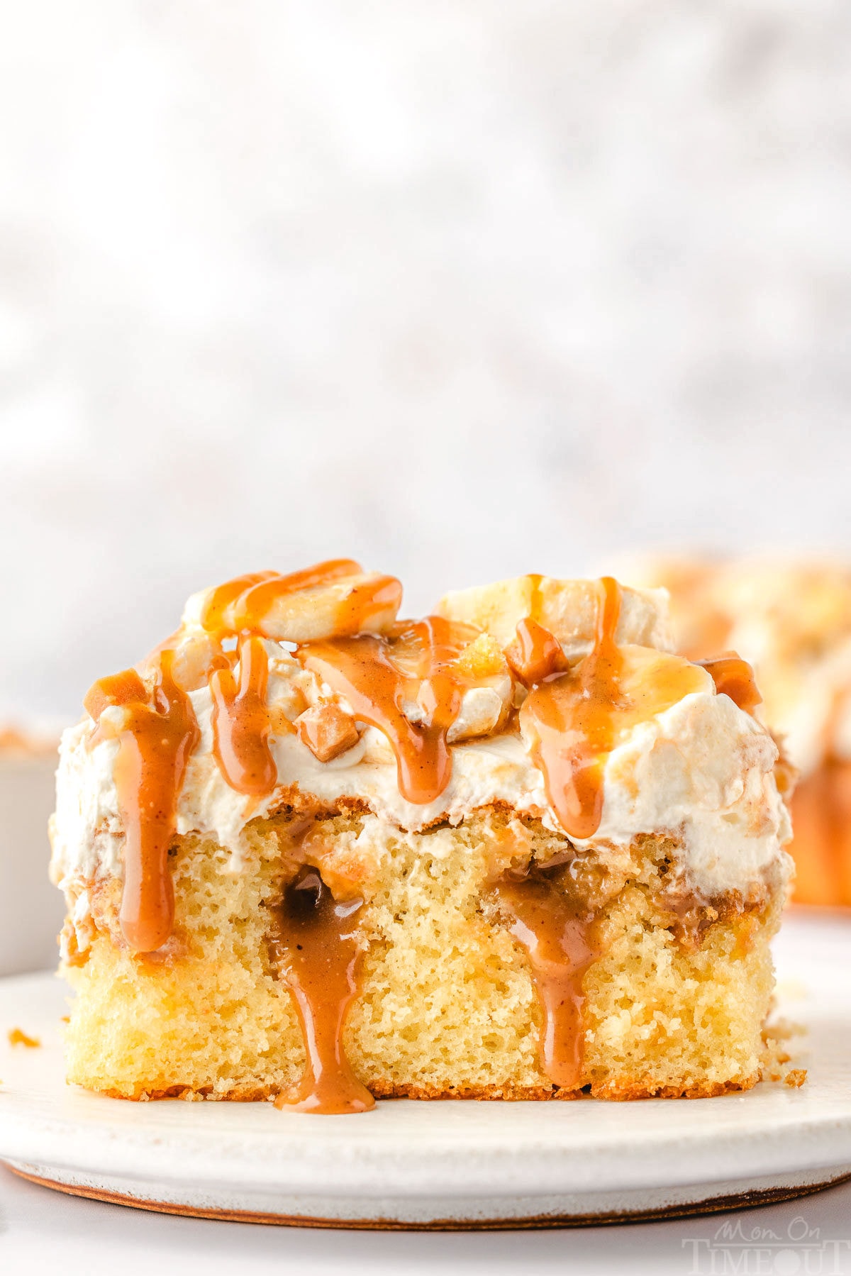 piece of banoffee poke cake on a white plate. the cake has been topped with whipped cream, bananas and dulce de leche sauce.