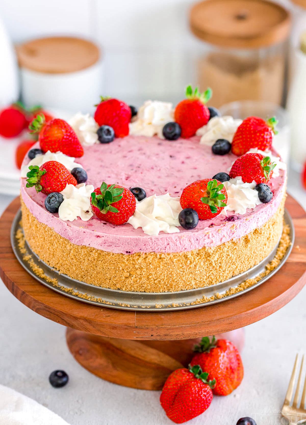 berry cheesecake topped with fresh blueberries and strawberries and whipped cream on a wood cake stand with more berries scattered around the base of the stand.