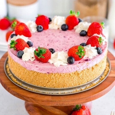 berry cheesecake topped with fresh blueberries and strawberries and whipped cream on a wood cake stand with more berries scattered around the base of the stand.
