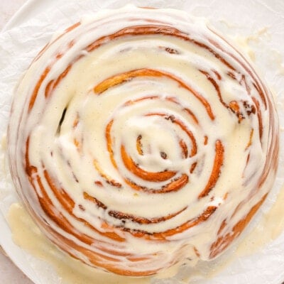 top down view of a giant cinnamon roll cake sitting on a white piece of parchment paper with a cream cheese glaze generously drizzled over the top.