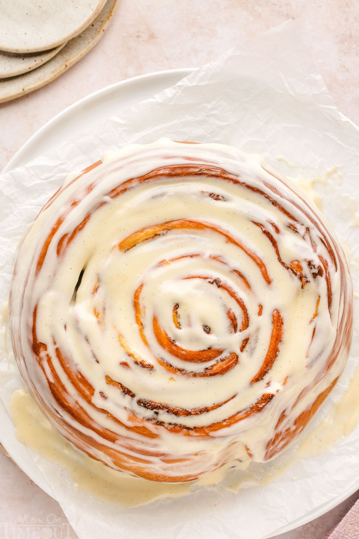 giant cinnamon roll with a cream cheese glaze sitting on white parchment with small plates to the side.