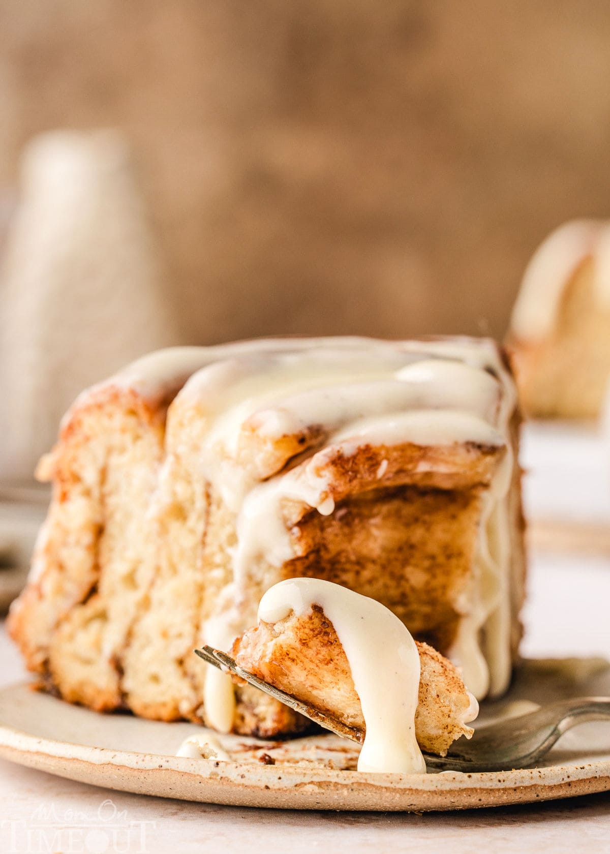 Slice of giant cinnamon roll cake recipe on small white plate. Cake has been drizzled with cream cheese glaze. A fork has taken the tip off the slice.