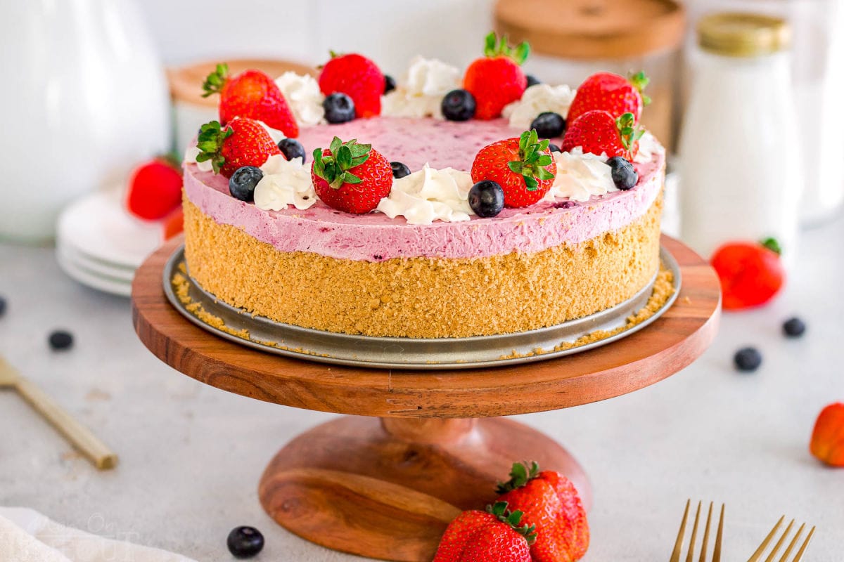 wide shot of berry cheesecake topped with fresh berries and whipped cream. Golden brown graham cracker crust can be seen. the cheesecake is on a wood cake stand.