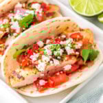 three chicken tacos on a white rectangular plate topped with pico de gallo, cilantro and queso fresco. A half a lime can be seen in the background.