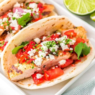 three chicken tacos on a white rectangular plate topped with pico de gallo, cilantro and queso fresco. A half a lime can be seen in the background.