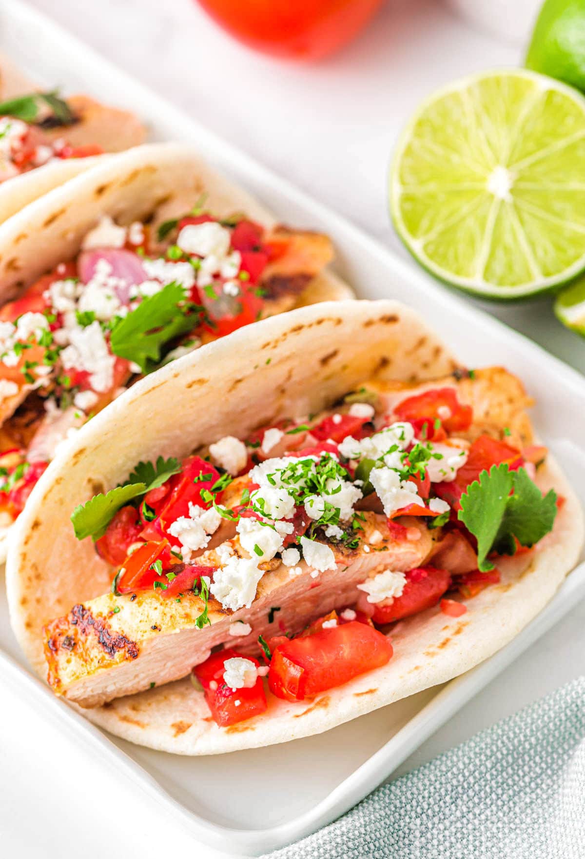 three chicken tacos on a white rectangular plate topped with pico de gallo, cilantro and queso fresco. A half a lime can be seen in the background.