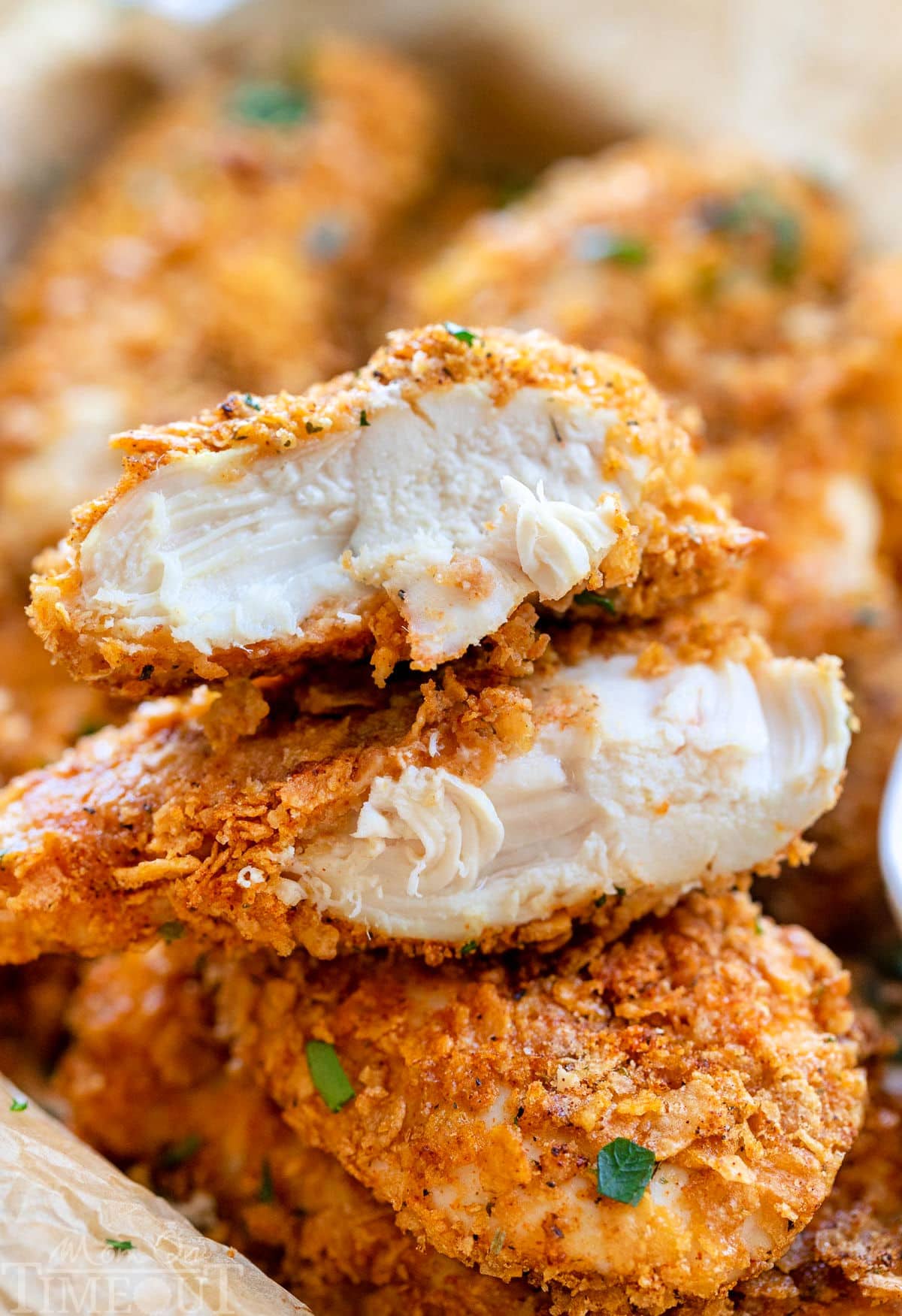 cornflake chicken in a pile with ranch dipping sauce. Top chicken tender is cut in half and stacked on each other showing the moist chicken and flavorful cornflake coating.