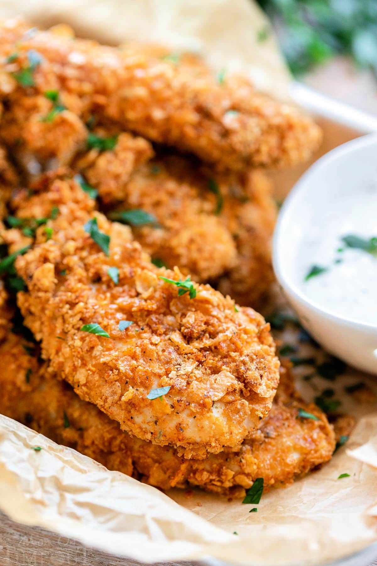 small baking sheet lined with brown parchment and piled high with cornflake chicken strips. a small bowl of ranch sits next to the chicken.