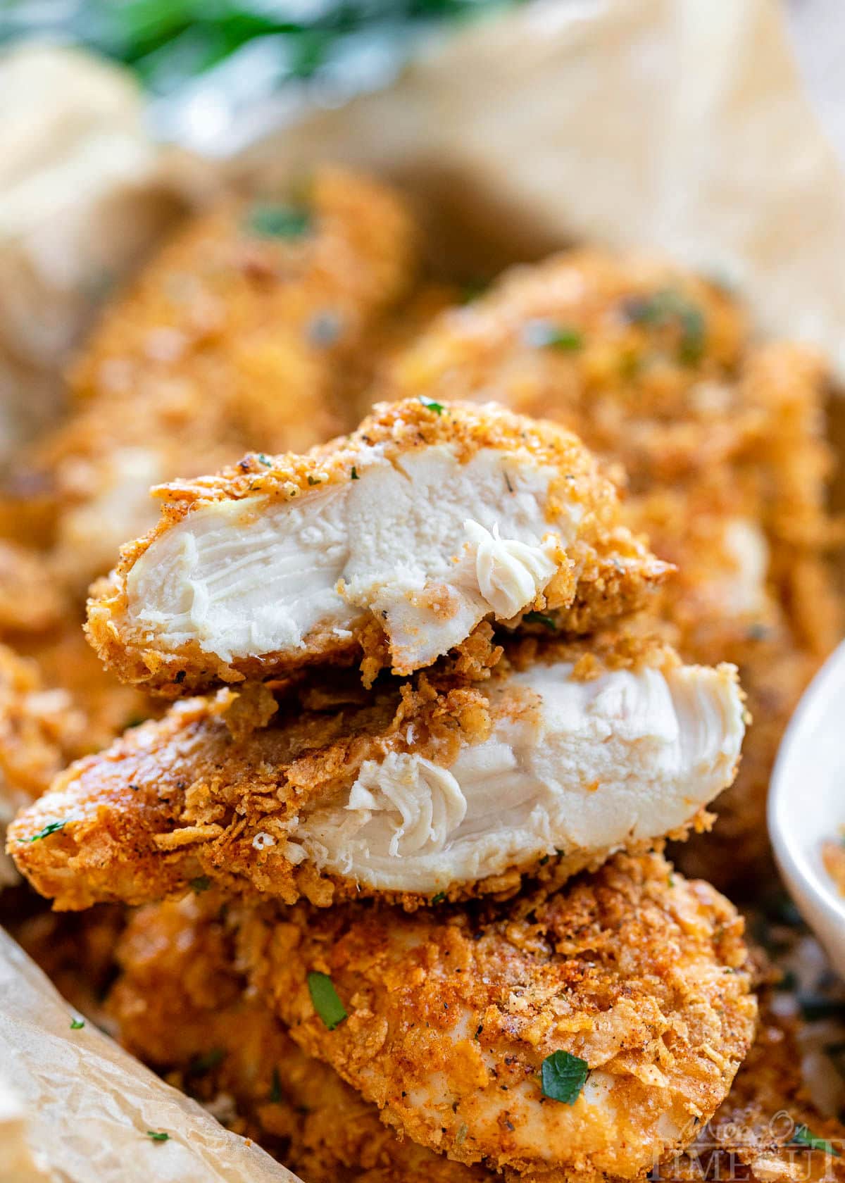 cornflake chicken in a pile with ranch dipping sauce. Top chicken tender is cut in half and stacked on each other showing the moist chicken and flavorful cornflake coating.