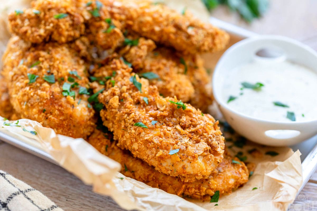 small baking sheet lined with brown parchment and piled high with cornflake chicken strips. a small bowl of ranch sits next to the chicken.