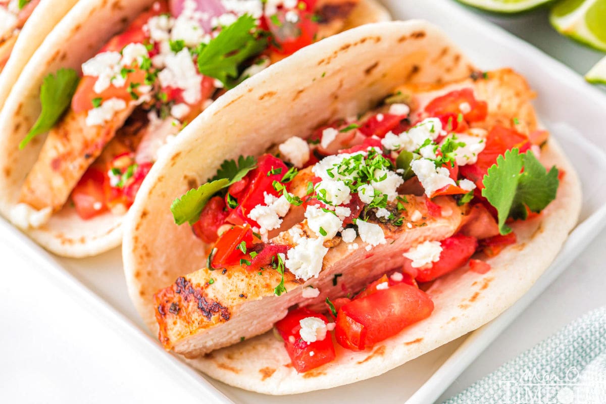 three chicken tacos on a white rectangular plate topped with pico de gallo, cilantro and queso fresco. A half a lime can be seen in the background.
