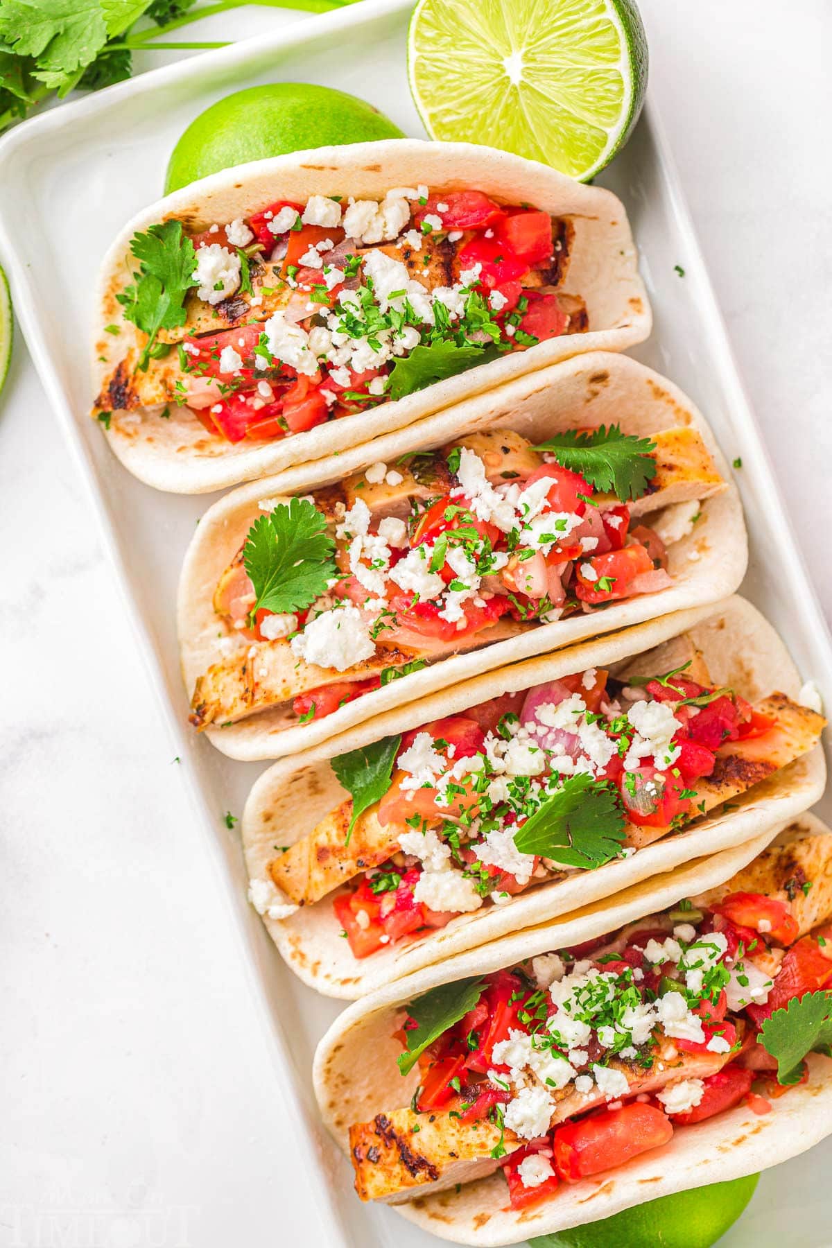 top down look at four grilled chicken tacos on a white plate topped with pico de gallo, cilantro and queso fresco. Lime wedges are scattered around the tacos.