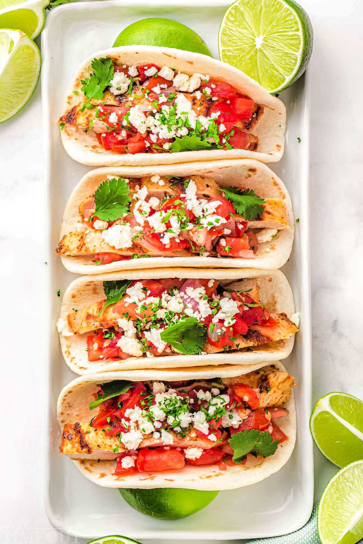 top down look at four grilled chicken tacos on a white plate topped with pico de gallo, cilantro and queso fresco. Lime wedges are scattered around the tacos.