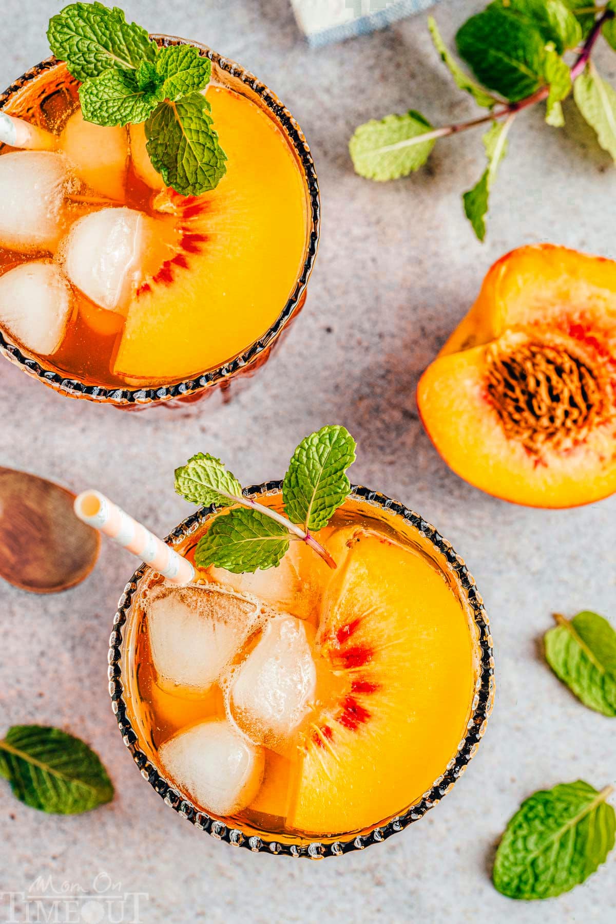 top down shot of two glasses of peach iced tea with mint and peach garnishes. both glasses have paper straws in them and half of a peach with a pit in it next to the glasses.