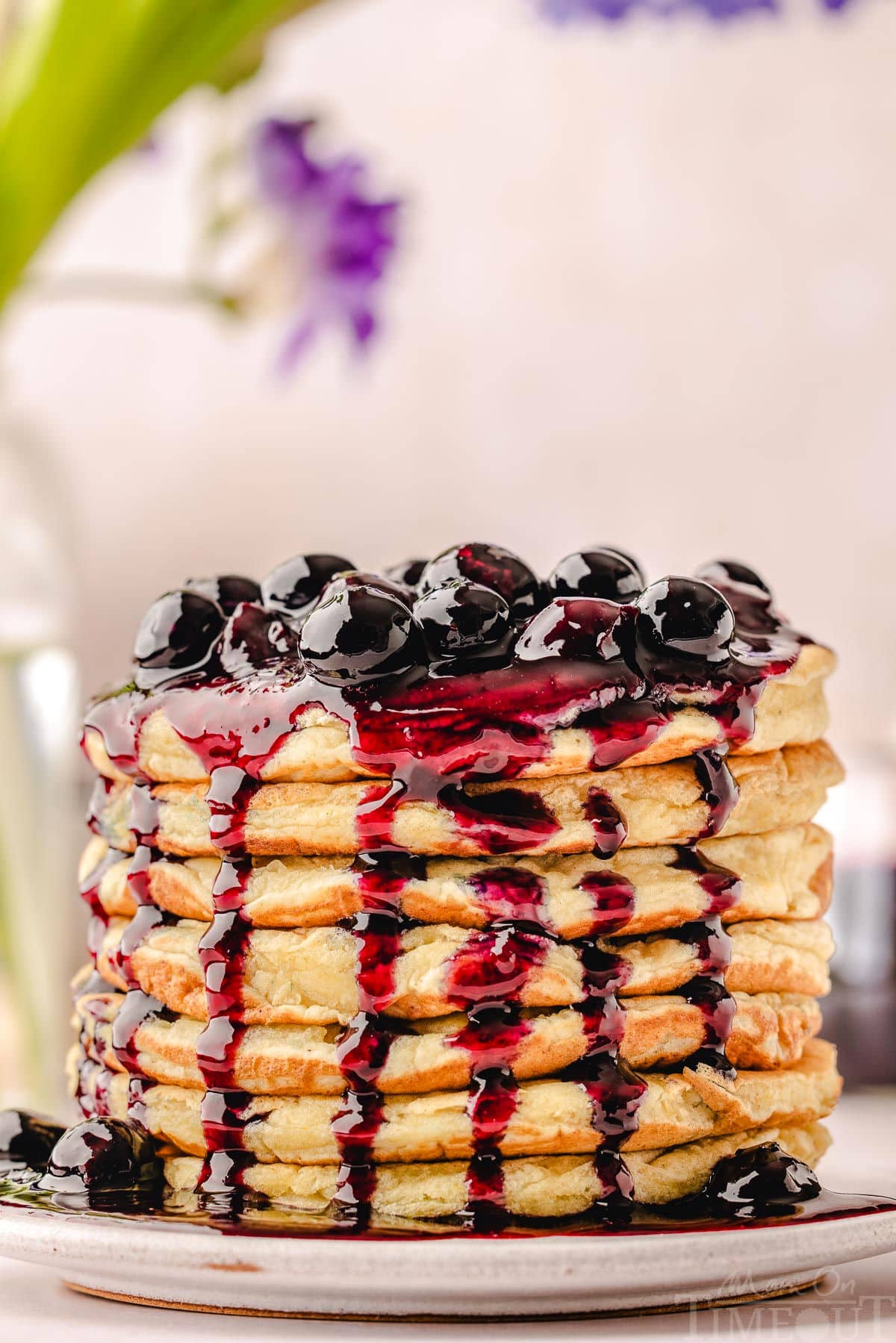 lemon ricotta waffles stacked high on a white round plate and topped with a glistening homemade blueberry sauce. fresh flowers can be seen in the background.