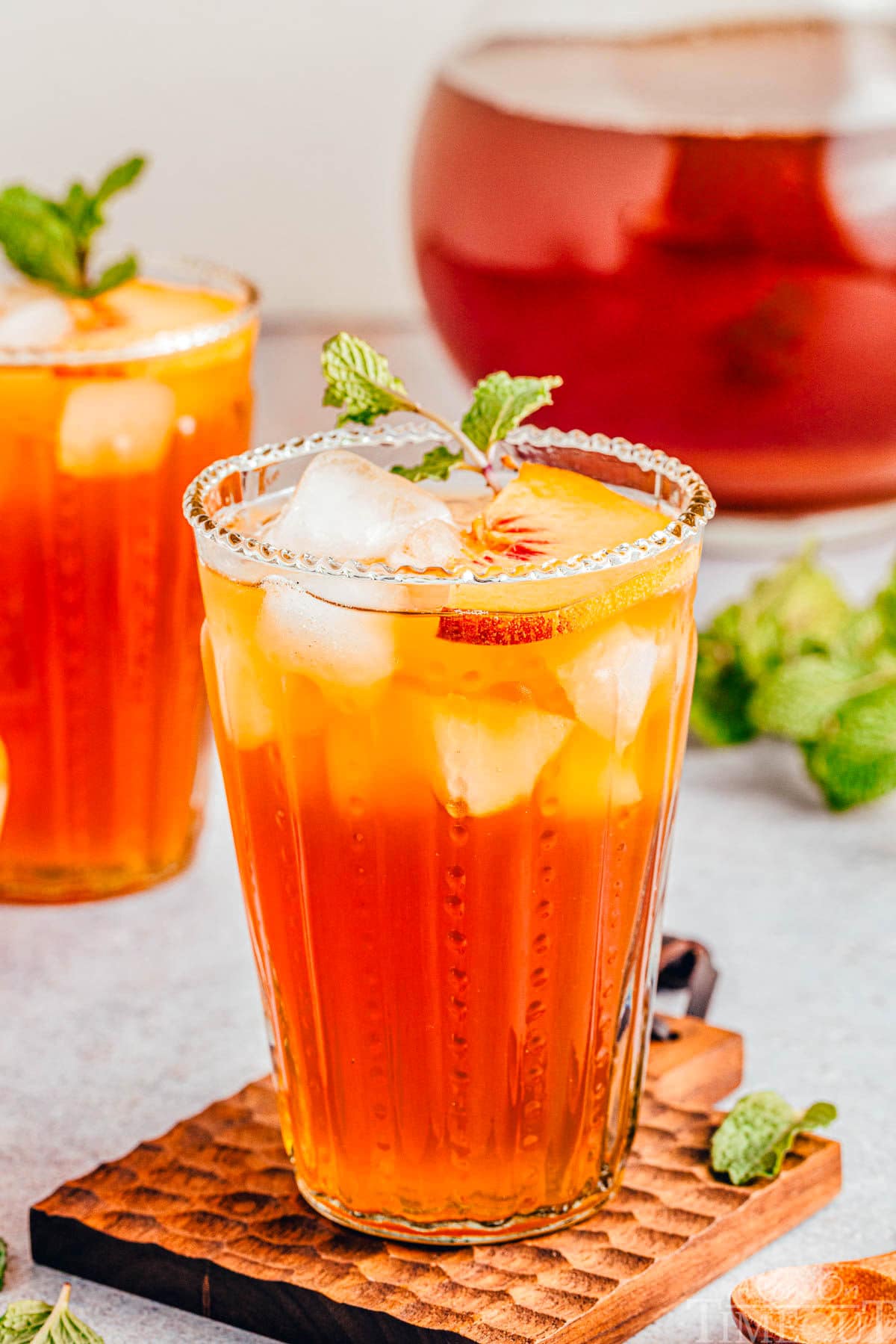 tall glass of refreshing peach tea sitting in front of a pitcher of tea and another glass.