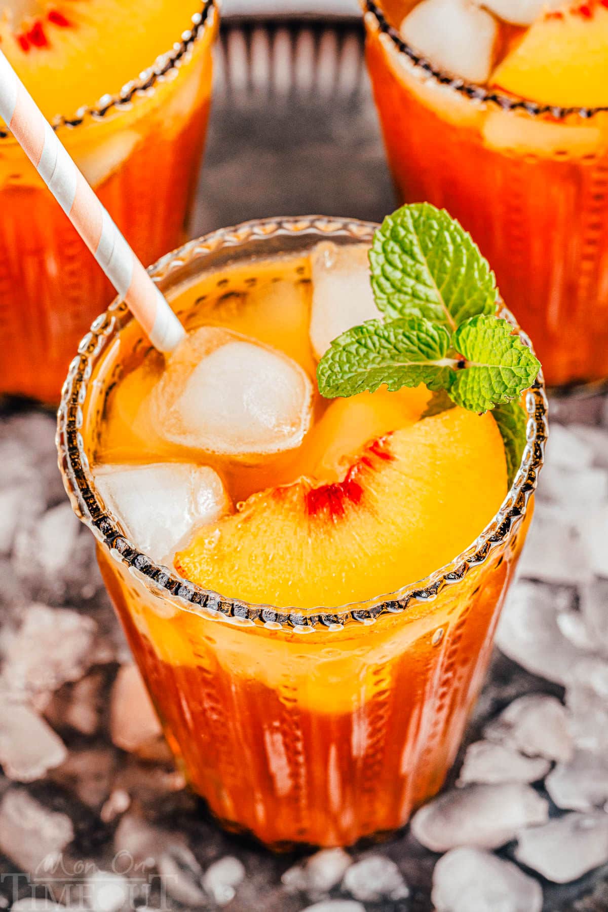 three tall glasses filled with homemade peach iced tea and topped with fresh peach slices and a sprig of mint. glasses are sitting on a tray covered with ice.