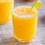 side view of a pineapple mango mocktail in a glass with a lime wedge garnish on the rim.