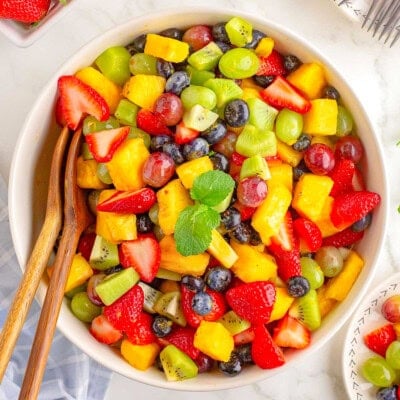 A top down view of fruit salad in a white bowl with wood salad tongs sticking out of it.