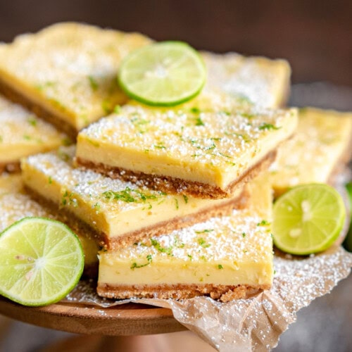wood cake stand piled high with key lime bars topped with key lime zest and a light dusting of powdered sugar. Slices of lime are scattered around the bars.