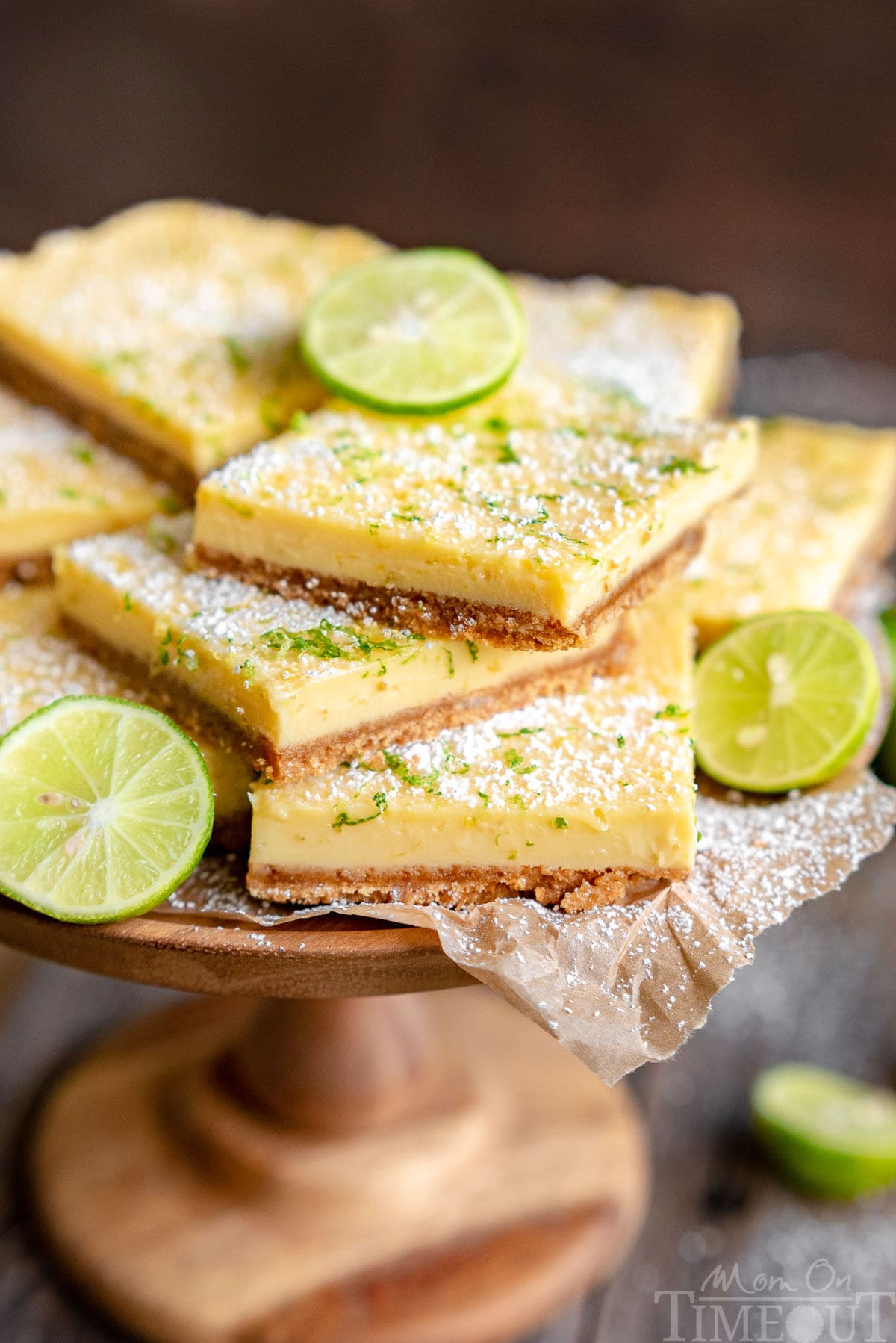 wood cake stand piled high with key lime bars topped with key lime zest and a light dusting of powdered sugar. Slices of lime are scattered around the bars.