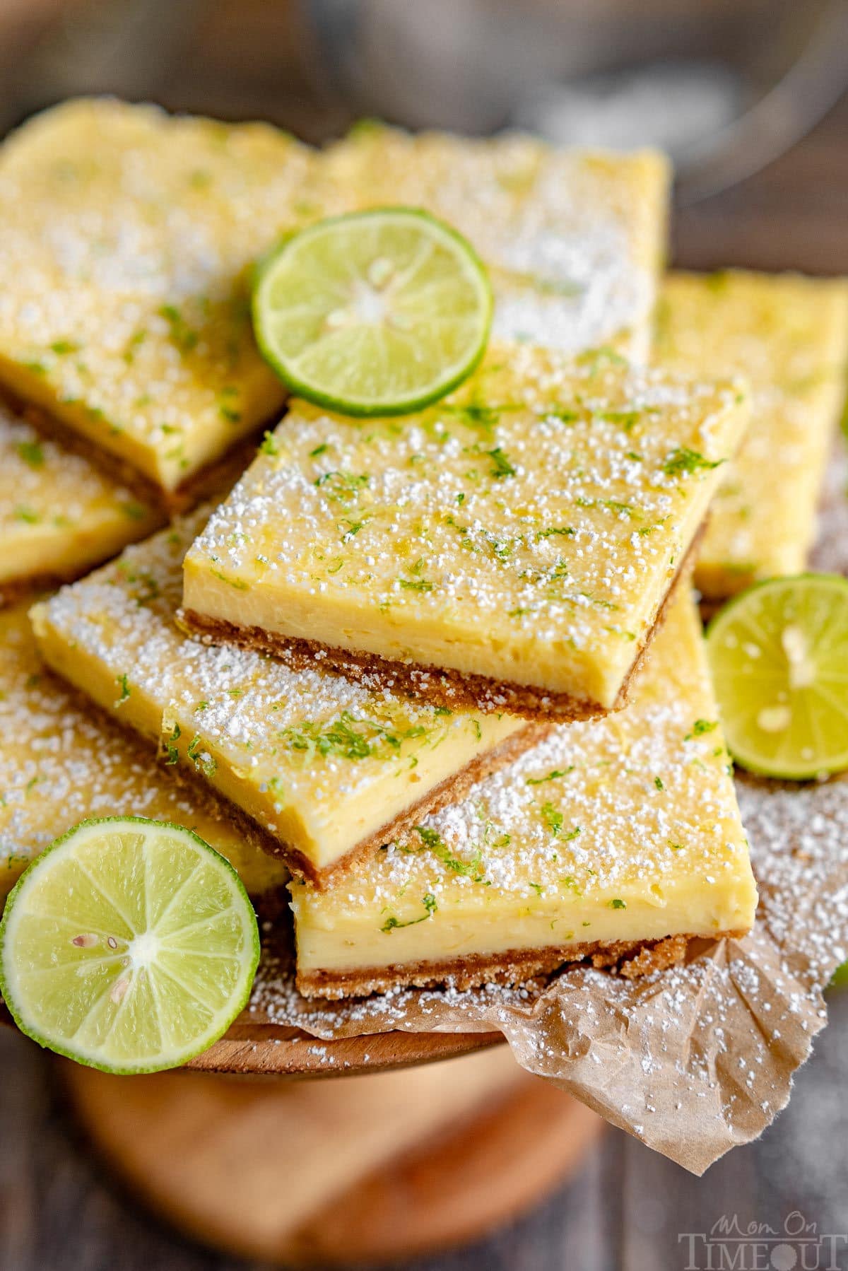close up look at key lime bars piled high on a wood cake stand lined with parchment paper. Bars have been topped with lemon and lime zest and powdered sugar.