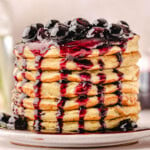 lemon ricotta waffles stacked high on a white round plate and topped with a glistening homemade blueberry sauce.