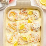 An overhead view of a pan of lemon sweet rolls topped with a lemon cream cheese glaze, lemon slices and lemon zest. Baking dish is white.