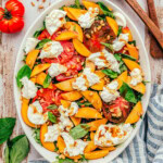 fresh peach salad made with burrata and heirloom tomatoes all topped with a balsamic vinaigrette. Salad is served on a white oval platter and is topped with pine nuts.