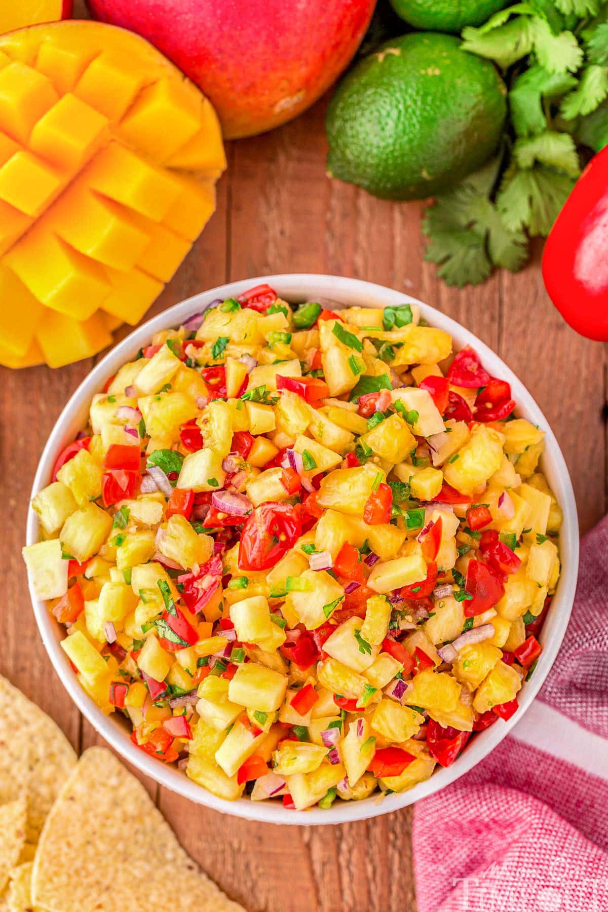 top down look at pineapple mango salsa in a white bowl surround by chips, mango, limes and cilantro. 
