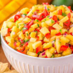 serving bowl filled withe pineapple mango salsa ready to be enjoyed. Mangoes can be seen in the background.