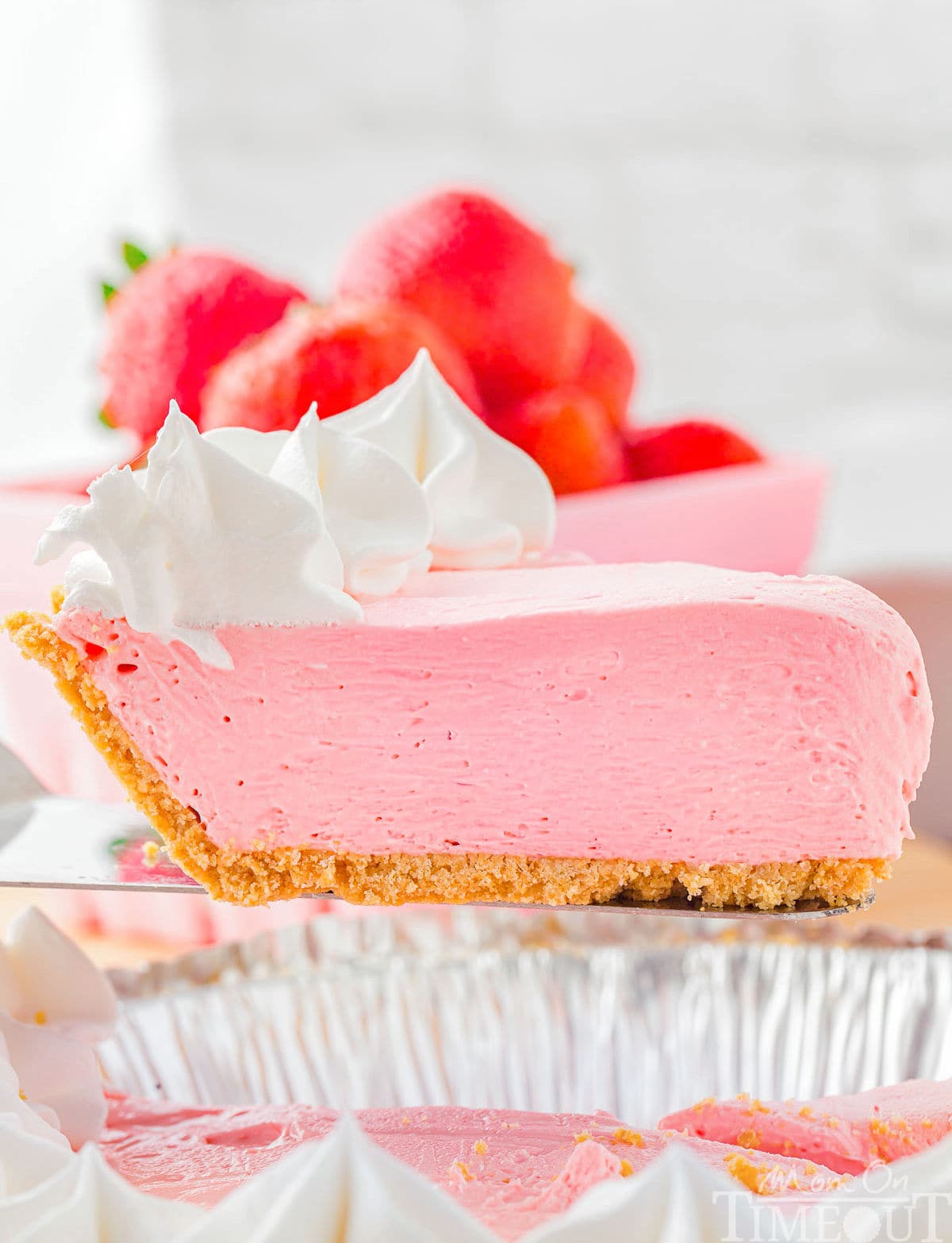 Pink slice of kool aid pie recipe topped with strawberries and whipped cream being held by a server over the rest of the kool aid pie.