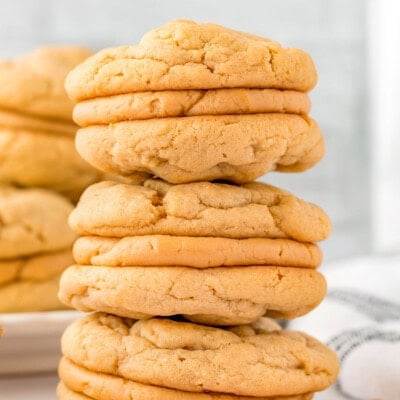 Three peanut butter sandwich cookies stacked on each other on a marble surface. A plate of more cookies can be seen in the background.