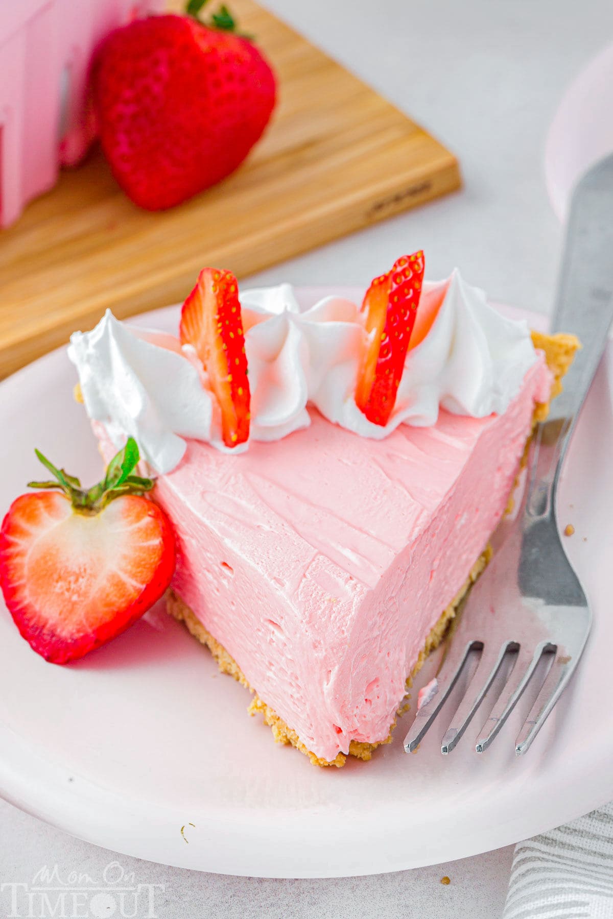 Slice of kool aid pie topped with whipped cream and strawberries on small round white plate with a fork sitting next to it.
