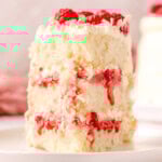 Slice of white chocolate raspberry cake on a white round plate topped with fresh raspberries and white chocolate curls.