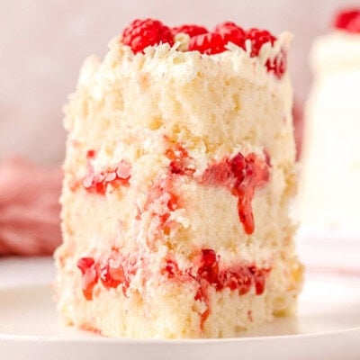 Slice of white chocolate raspberry cake on a white round plate topped with fresh raspberries and white chocolate curls.