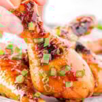 Honey glazed chicken drumstick garnished with green onions being picked up off a small sheet tray with more chicken on it.